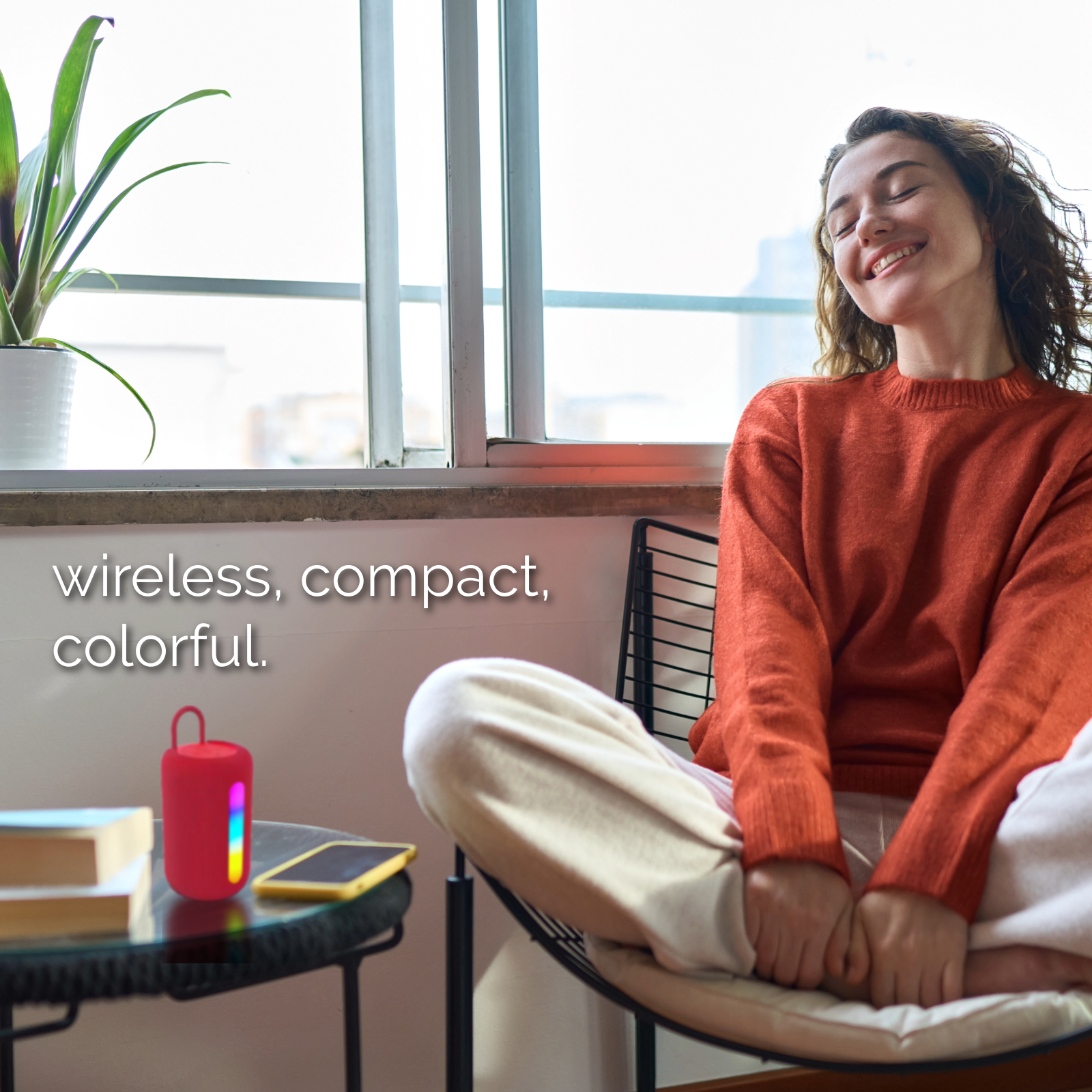Woman relaxing by the window with a red portable Bluetooth speaker on a table beside her.